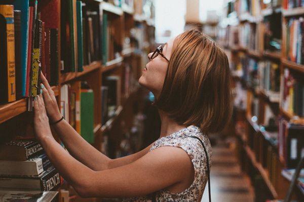 Element ilustracyjny: Młoda kobieta w okularach szukająca książki wśród bibliotecznych regałów